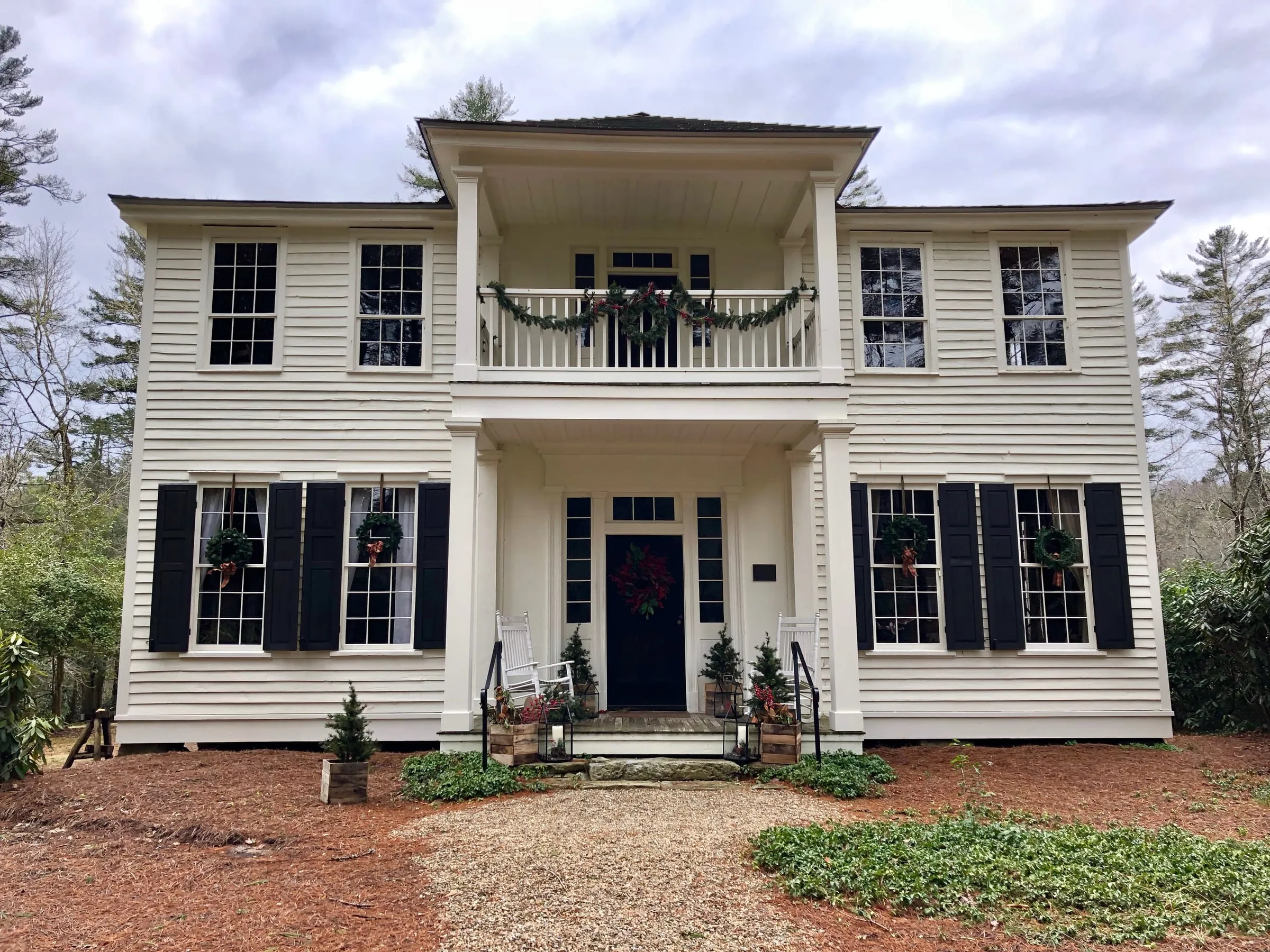 Two-story white traditional home with black shutters and a columned porch on a wooded lot.